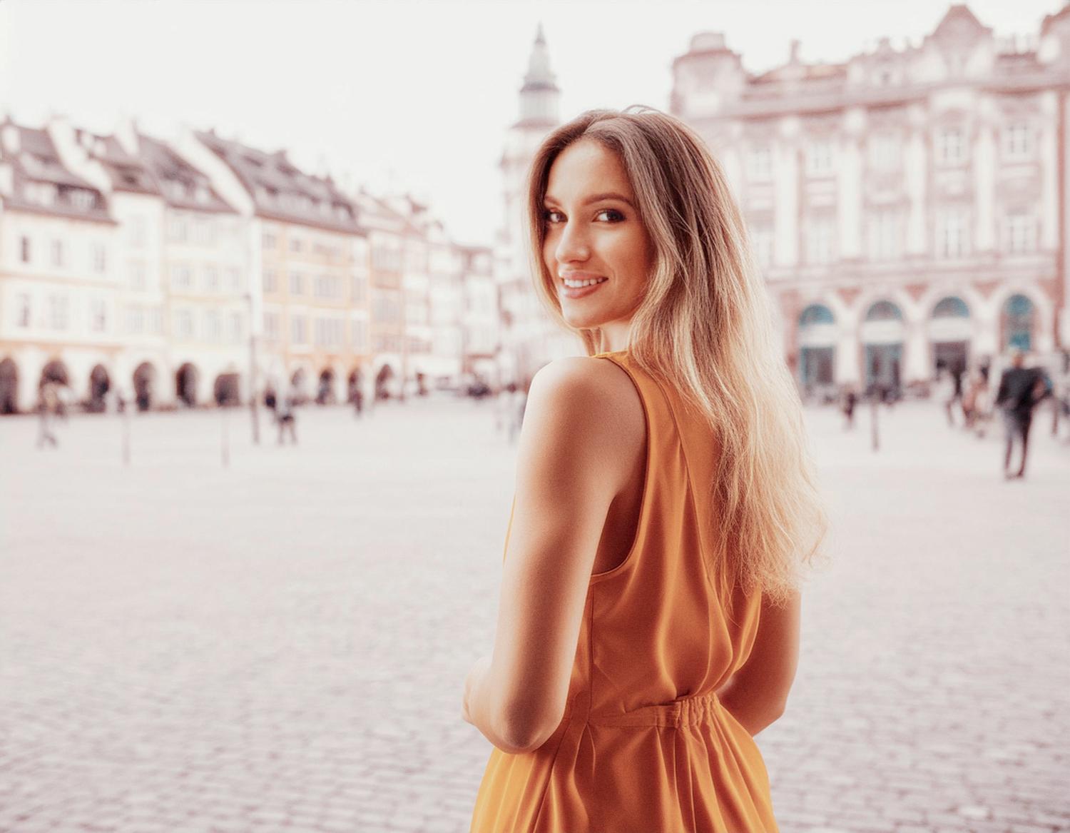 A joyful woman in an orange sleeveless dress glances at the camera, exuding chic style amidst a European town square's historic charm.