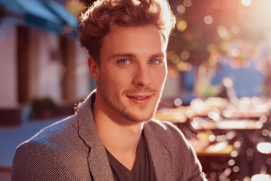A young man with wavy blonde hair and a light beard smiles warmly, sporting a chic gray blazer from the Travel Line over a dark V-neck. He stands before a sunlit outdoor cafe, exuding effortless style.