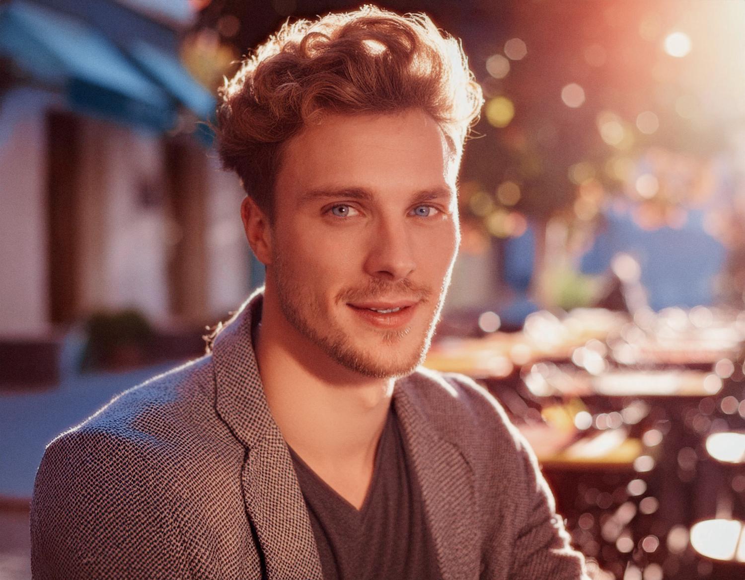 A young man with wavy blonde hair and a light beard smiles warmly, sporting a chic gray blazer from the Travel Line over a dark V-neck. He stands before a sunlit outdoor cafe, exuding effortless style.