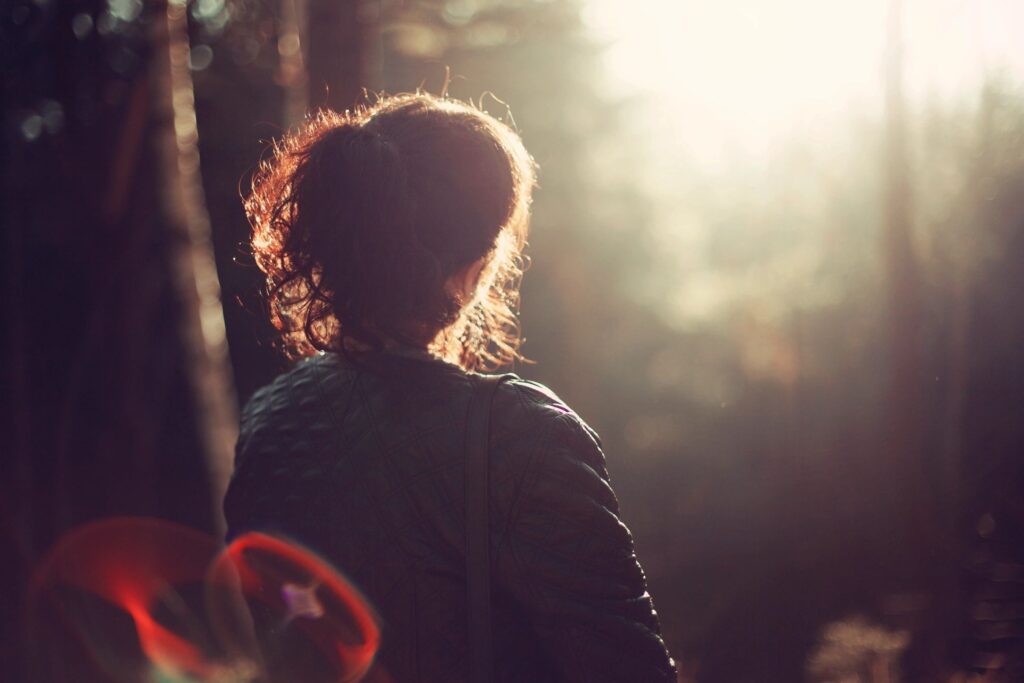 A curly-haired individual in a chic jacket stands amidst a forest, bathed in enchanting sunlight and lens flares, epitomizing outdoor elegance.