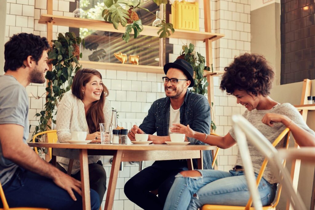 Four friends in a cozy café, laughing over coffee. White tile wall with stylish plant displays. Warm, inviting vibe perfect for casual chic moments.