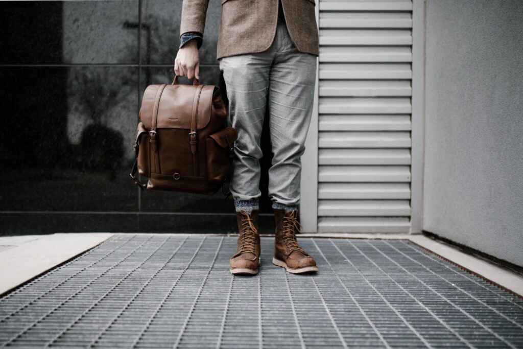 A stylish individual strikes a pose on a metal grate, sporting a brown leather backpack, blazer, and lace-up boots paired with grey checkered pants. Trendy urban backdrop.