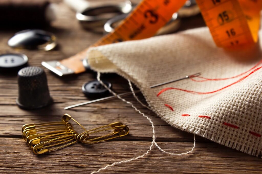 A close-up of a sewing workspace featuring fabric, a thimble, measuring tape, red-threaded needle, safety pins, buttons—crafting chic fashion pieces.