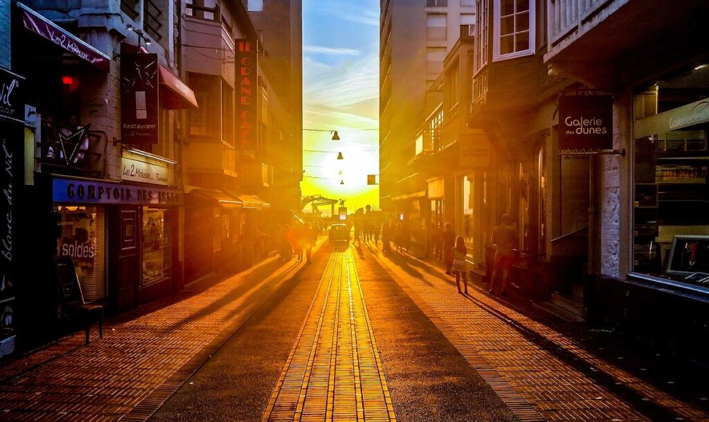 A narrow city street bathed in golden sunset hues, with shadows stretching along cobblestones. Shop signs peak out, and stylish silhouettes of pedestrians add charm.