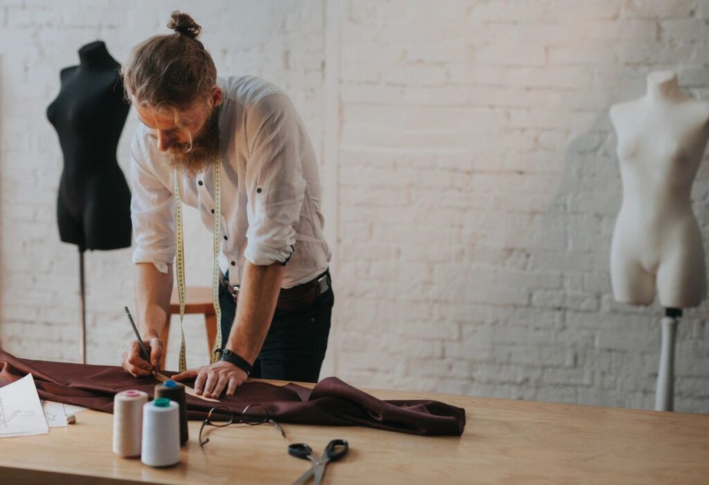 A bearded man with a long ponytail meticulously measures brown fabric on a wooden table. Dressed in a white shirt, he's surrounded by scissors, thread spools, and dress forms in a chic, brick-walled studio—capturing the essence of artisanal fashion craftsmanship.