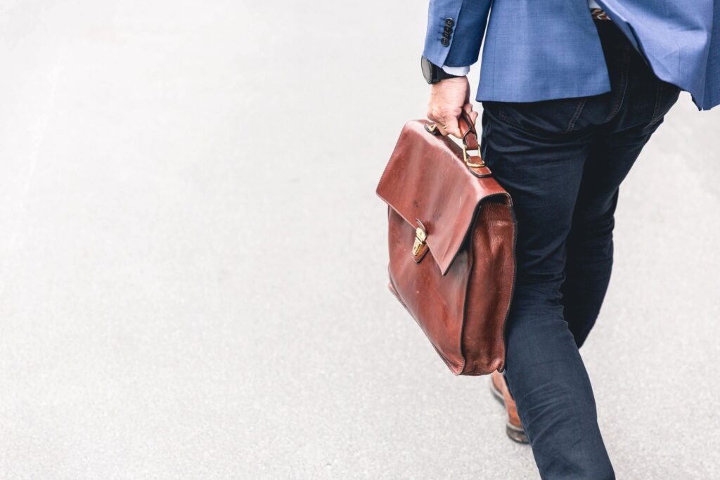 A stylish individual strides confidently on a grey path, showcasing a chic blue blazer and dark pants duo, accentuated by an elegant brown leather briefcase.