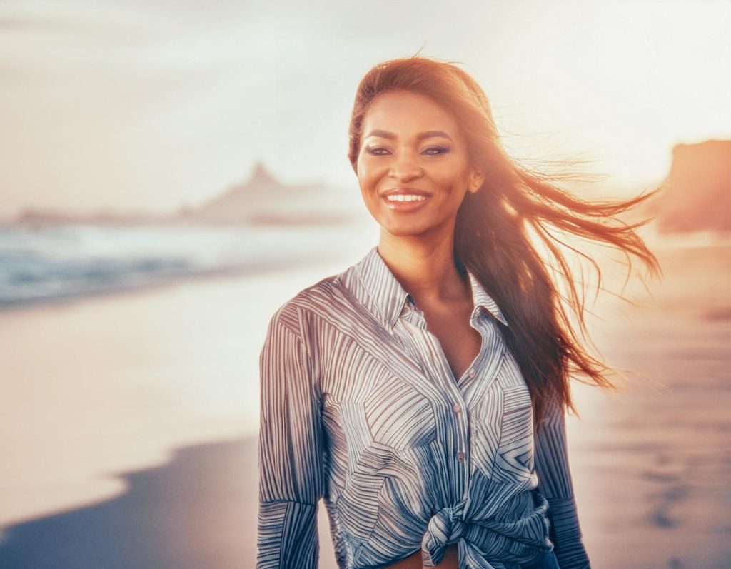 A joyful woman with long hair stands against a sunset beach backdrop, sporting a chic striped shirt tied at the waist and an Osprey Packs Heritage Musette.