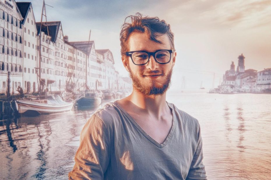 A bearded man with trendy glasses smiles by a serene waterfront at sunset. Boats and historic buildings reflect in the warm orange-blue sky—a stylish, picturesque moment.