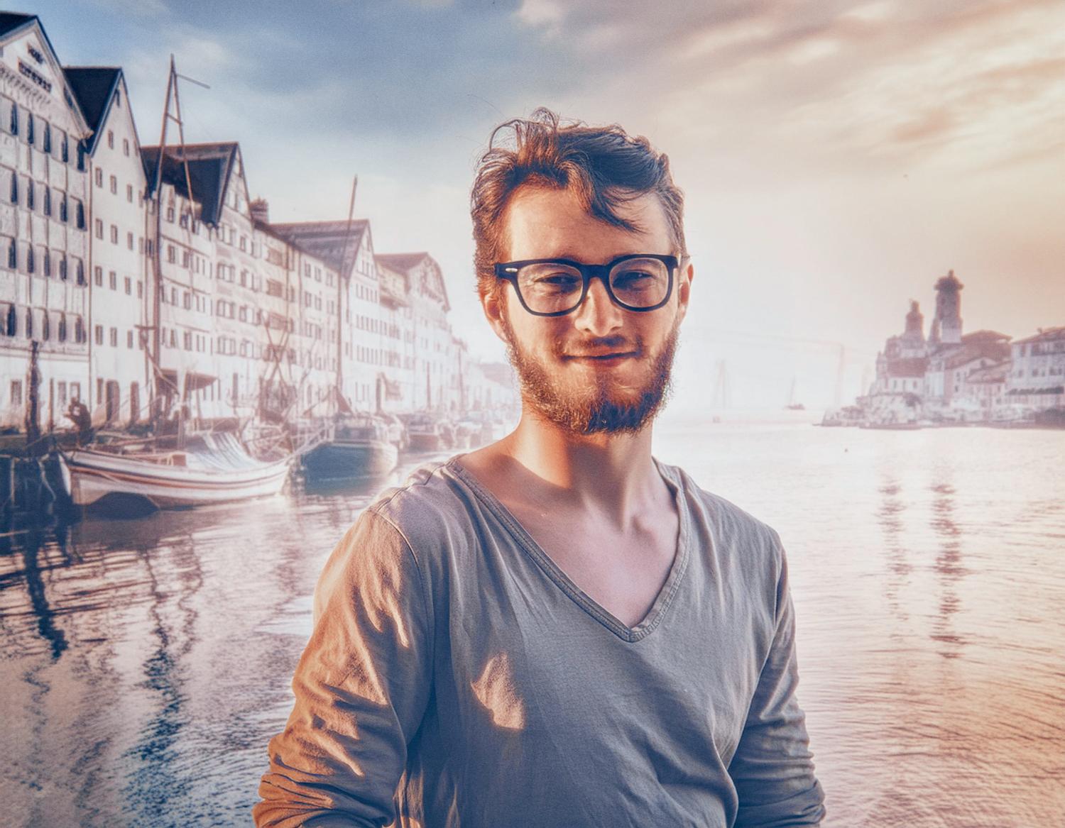 A bearded man with trendy glasses smiles by a serene waterfront at sunset. Boats and historic buildings reflect in the warm orange-blue sky—a stylish, picturesque moment.