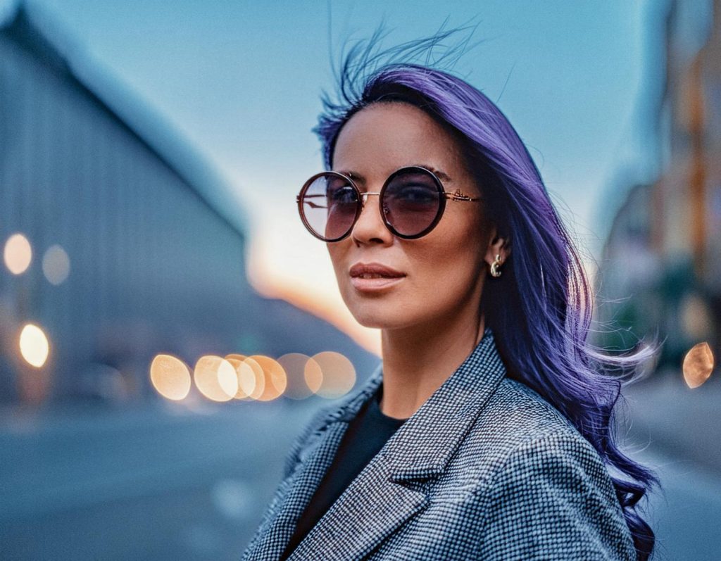 A trendsetter with long, purple hair gazes confidently into the camera, donning chic round sunglasses and a gray plaid jacket against a dusk cityscape backdrop.