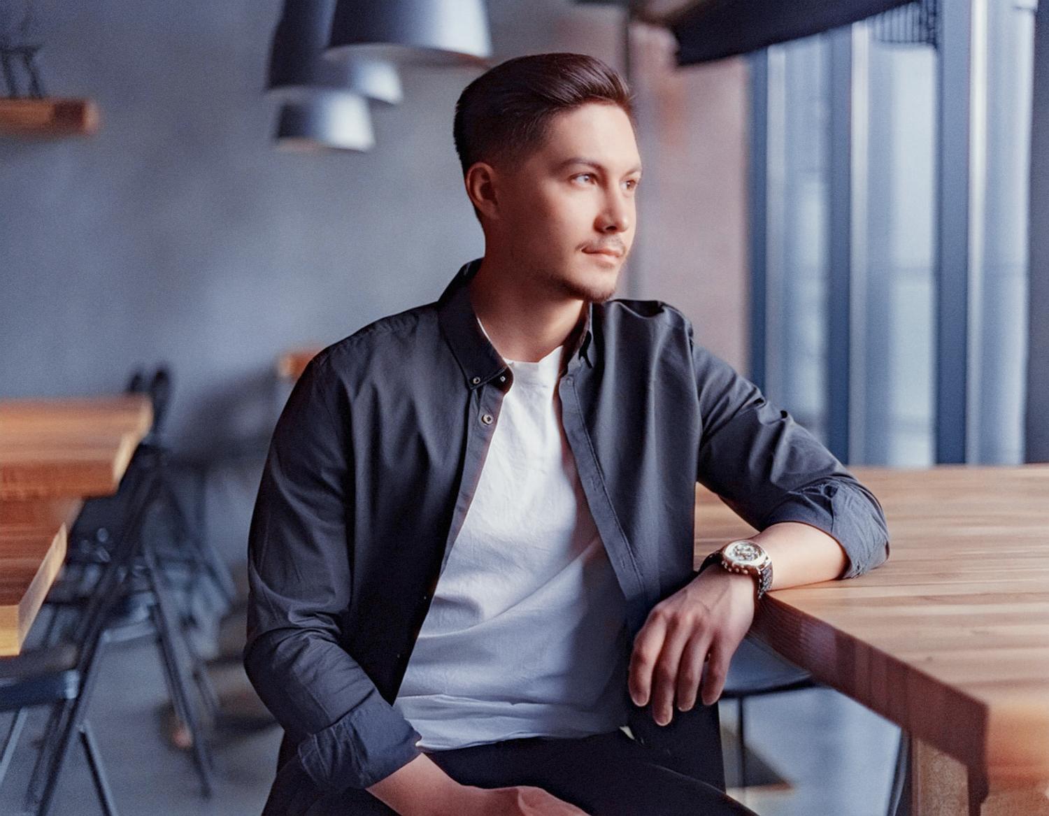 A stylish young man in a white shirt and dark jacket gazes thoughtfully out of a modern, minimalist cafe window. Industrial chic vibes.