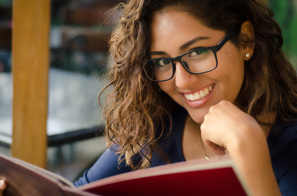 Curly-haired individual exudes style, sporting chic glasses and earrings, embracing a navy top by the window with an open book delight.
