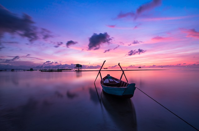 A flip-flop wearing someone sits on the shore at sunset beside a boat.