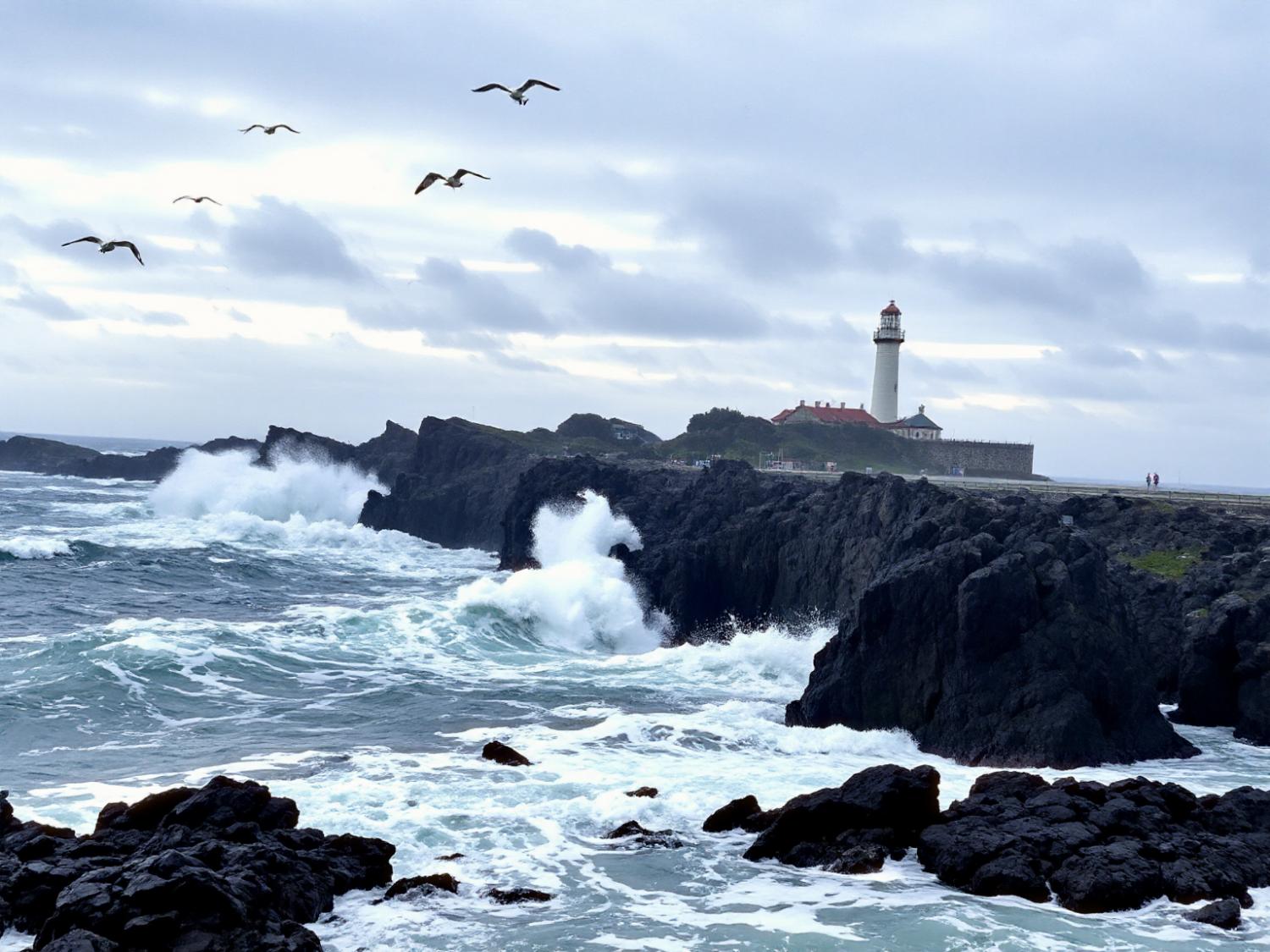 Amid crashing waves and rugged cliffs, two figures stroll by a lighthouse, capturing the coastal chic vibe on their Leia Lume Pad 2.