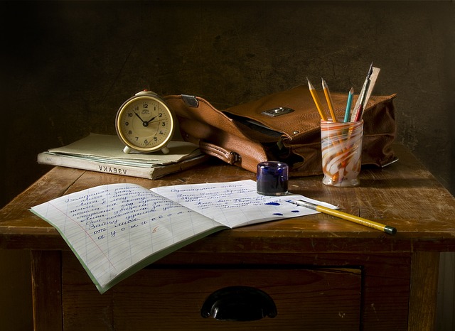 A vintage wooden desk with an open notebook, ink blots, pens in a colorful holder, an ink bottle, old-fashioned clock, and leather satchel exudes timeless style.