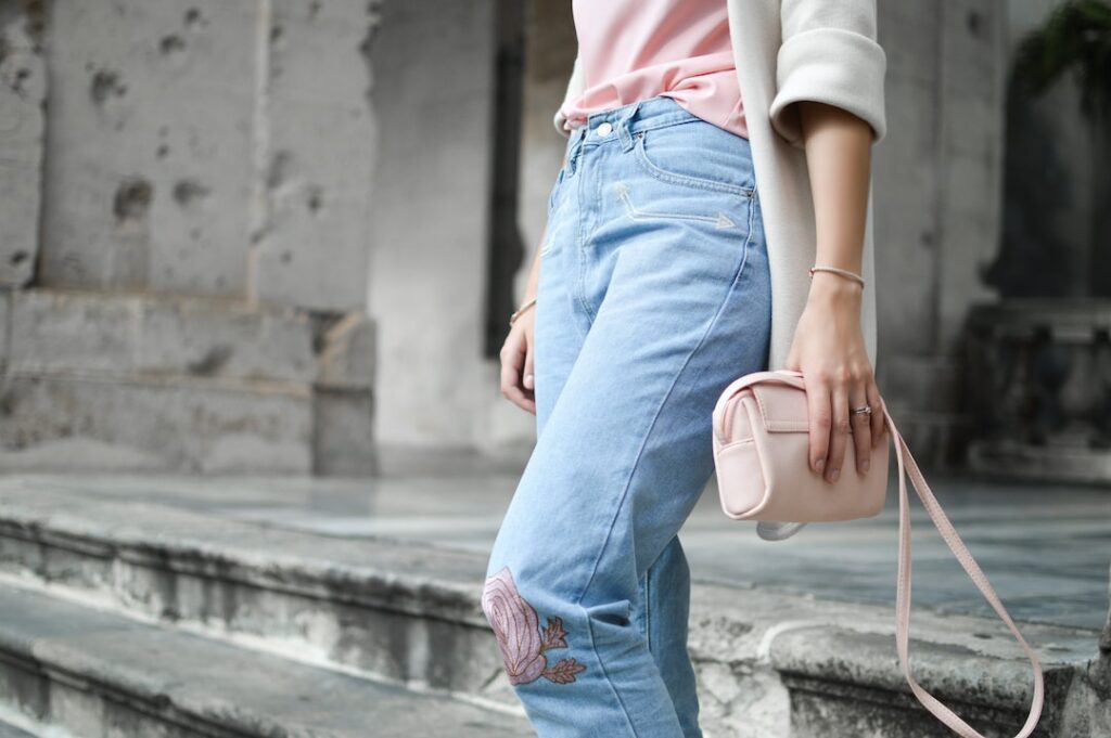 A stylish individual on stone steps, flaunting light blue jeans with pink rose embroidery, a chic pink top, and a white jacket draped over one shoulder. They accessorize with a small pink handbag and a ring on their left-hand ring finger. Perfect blend of elegance and casual flair.