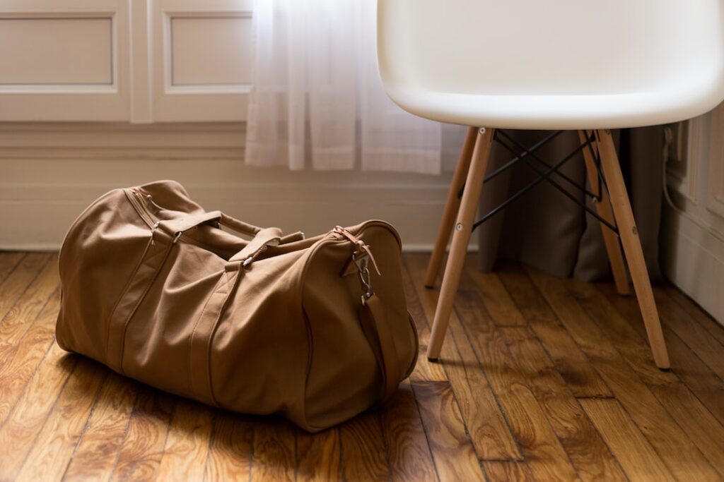 A chic tan canvas duffel bag rests stylishly on a wooden floor beside a sleek white chair. Sunlight filters in, casting a cozy, inviting glow.