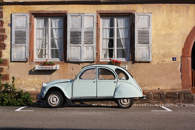 A light blue vintage car complements an old building's weathered yellow facade, adorned with stylish blue shutters and vibrant red flower boxes—a timeless fashion statement in architecture.