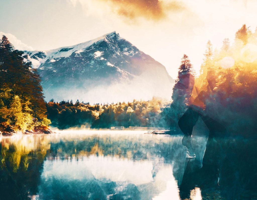 A serene sunrise over snow-capped peaks and a clear lake; golden light filters through trees, revealing a silhouette with stylish Filson Tin Cloth Bags.