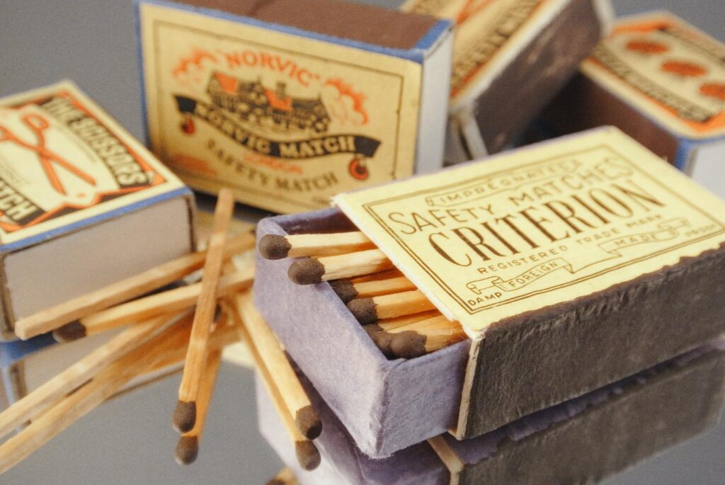 Vintage matchboxes with wooden dark-tipped matches showcase retro designs and brands like "Criterion" and "Norvic." A chic touch to any classic trench coat decor.