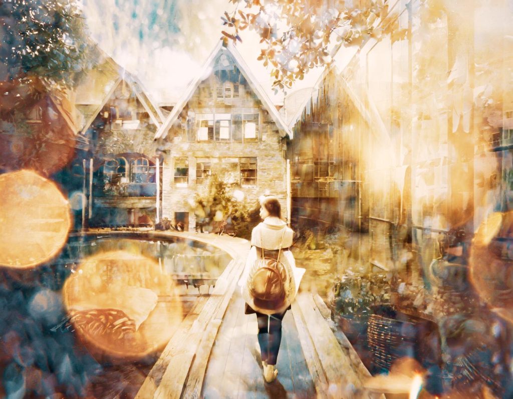 A stylish wanderer with a chic backpack and retro polarized sunglasses strolls a wooden path, flanked by dreamy bokeh and a stone building.