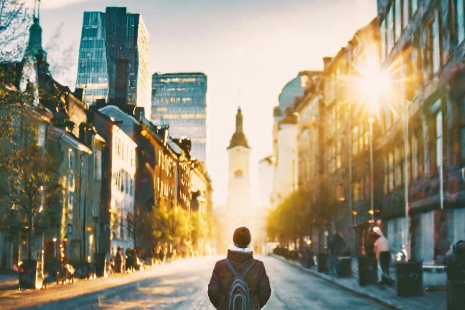 Urban chic: A stylish individual sports a beanie, backpack, and gleaming canvas belt buckle, standing amid sunlit streets and towering architecture.