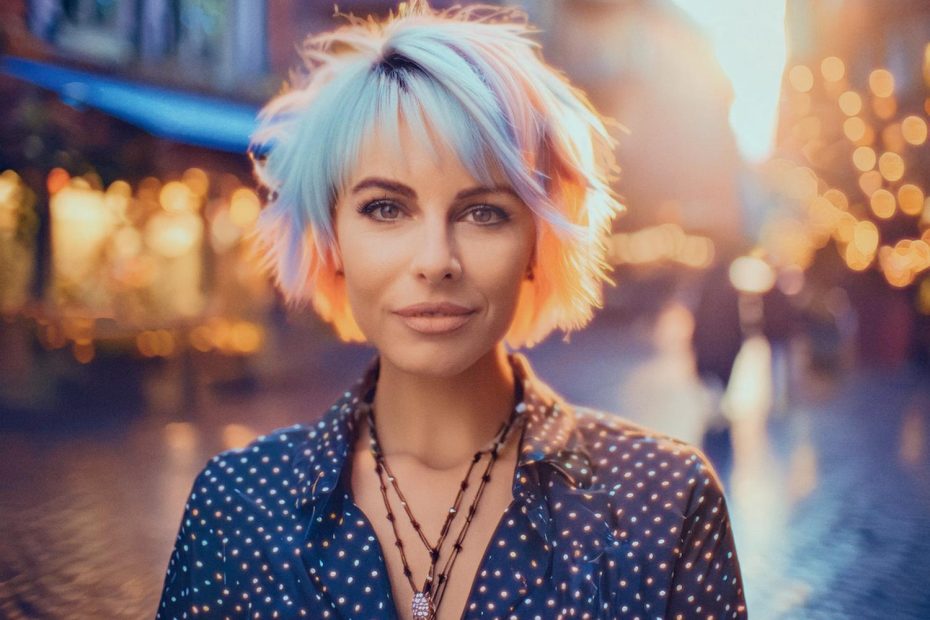 A trendsetter with pastel short hair and a polka dot blouse confidently shows off a blue messenger bag, set against a dreamy sunset bokeh backdrop.