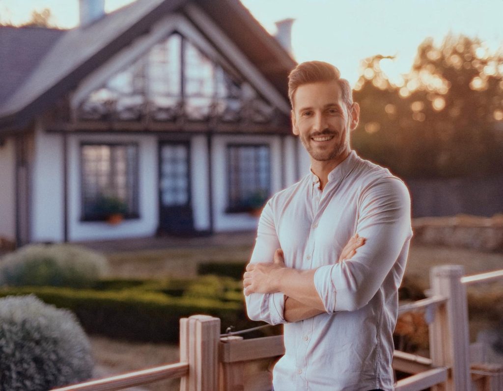 A confident man with a beard, sporting a light long-sleeve shirt and holding a chic brown leather messenger bag, stands before a charming house with a pitched roof and wooden balcony; lush garden backdrop.
