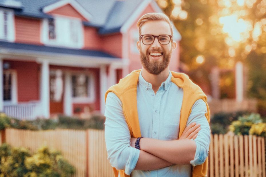 A bearded man with glasses radiates style in a light blue shirt and yellow sweater over his shoulders, rocking a perfect messenger bag. He stands in front of a charming red house with a white porch and picket fence, bathed in sunlight filtering through lush greenery.