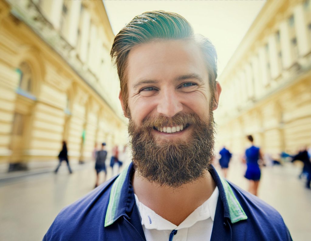 A bearded man radiates style on a sunny city street, pairing a crisp white shirt with a blue jacket. Yellow buildings and passersby frame his fashionable look, perfect for flaunting an Ita Messenger Bag.