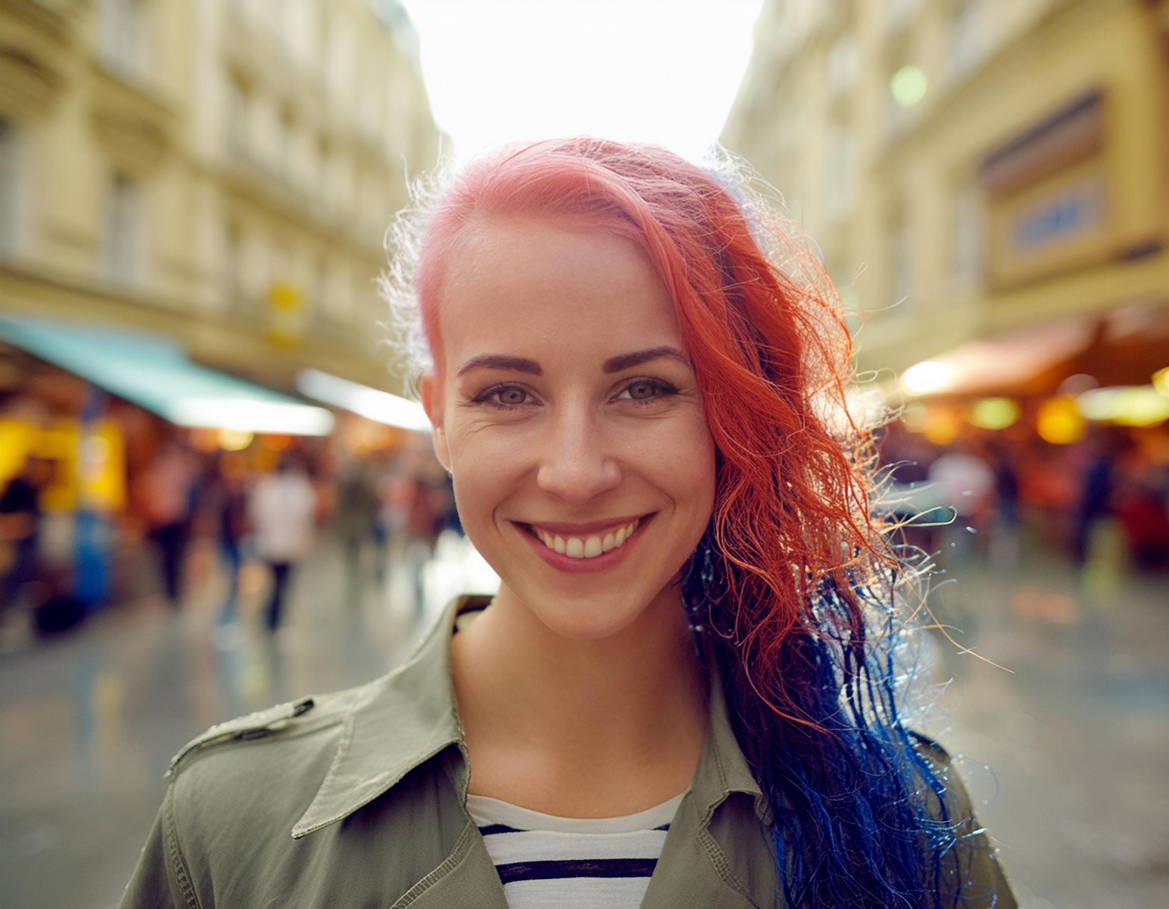 A person with vibrant red and blue hair beams confidently while standing on a lively street. They rock an Ita Messenger Bag, blending fashion with personal flair.