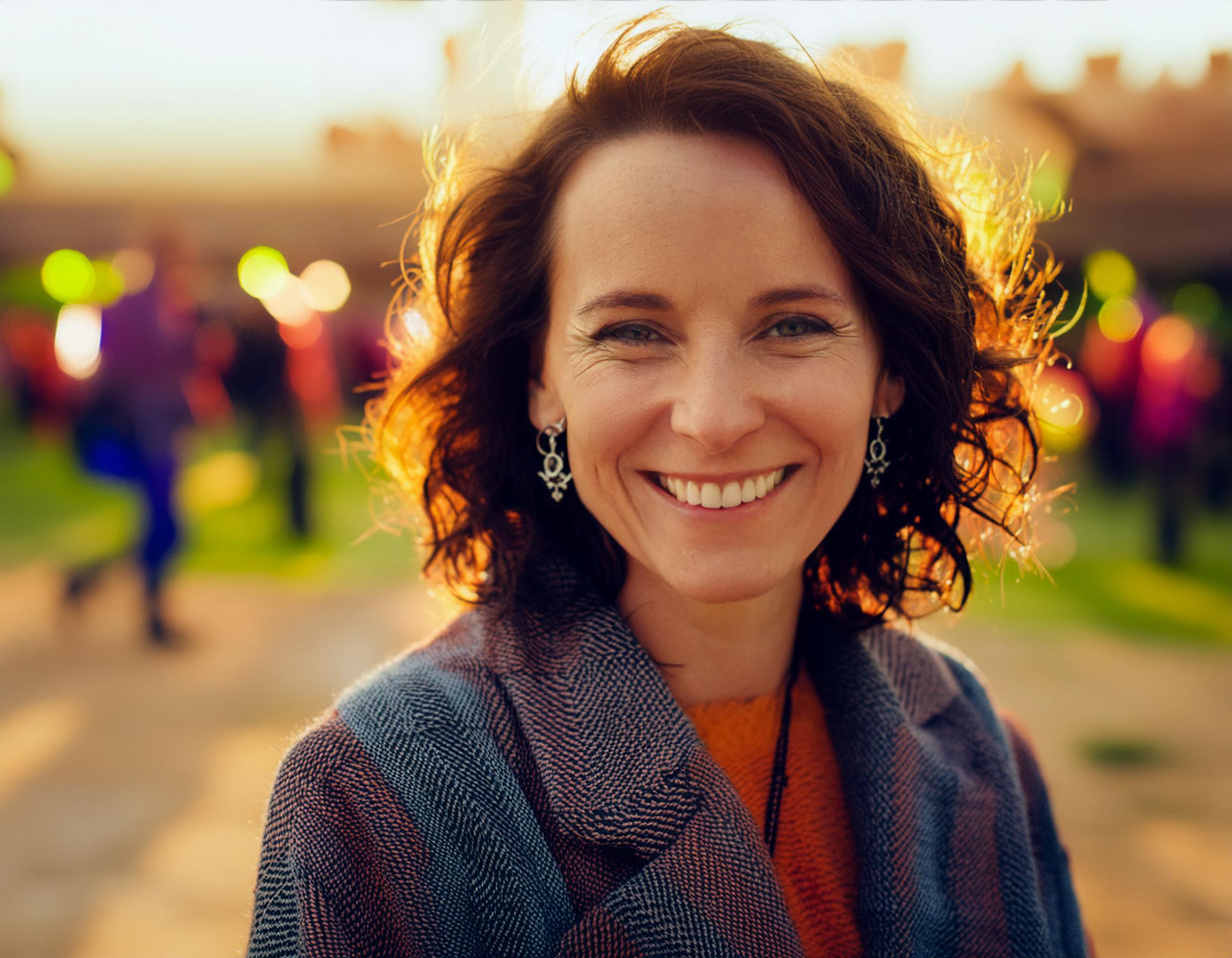 A woman with curly hair beams, wearing a chic textured coat. Her vibrant Ita Messenger Bag stands out at a colorful sunset festival, merging personal flair with current fashion trends.