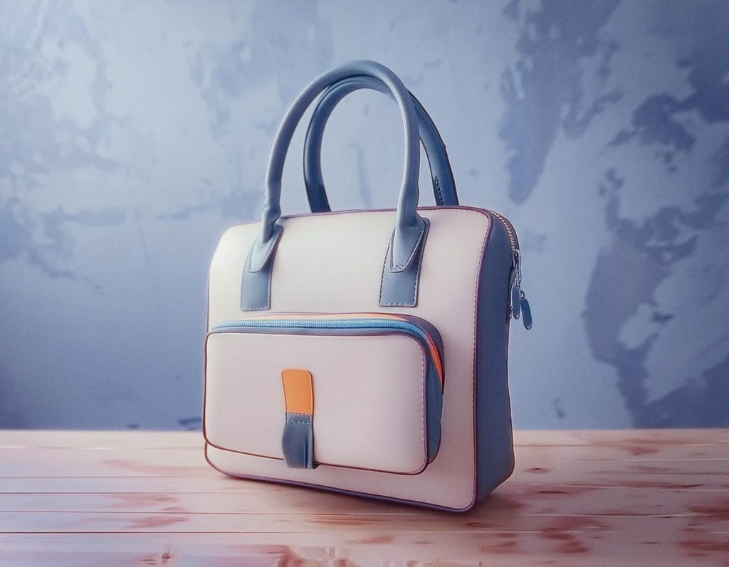 A chic shoulder bag showcases a white body, light blue handles, and trim, with an orange-accented front pocket. Perfectly styled on a wooden surface against a marbled blue-grey backdrop. Fashion-forward accessory alert!