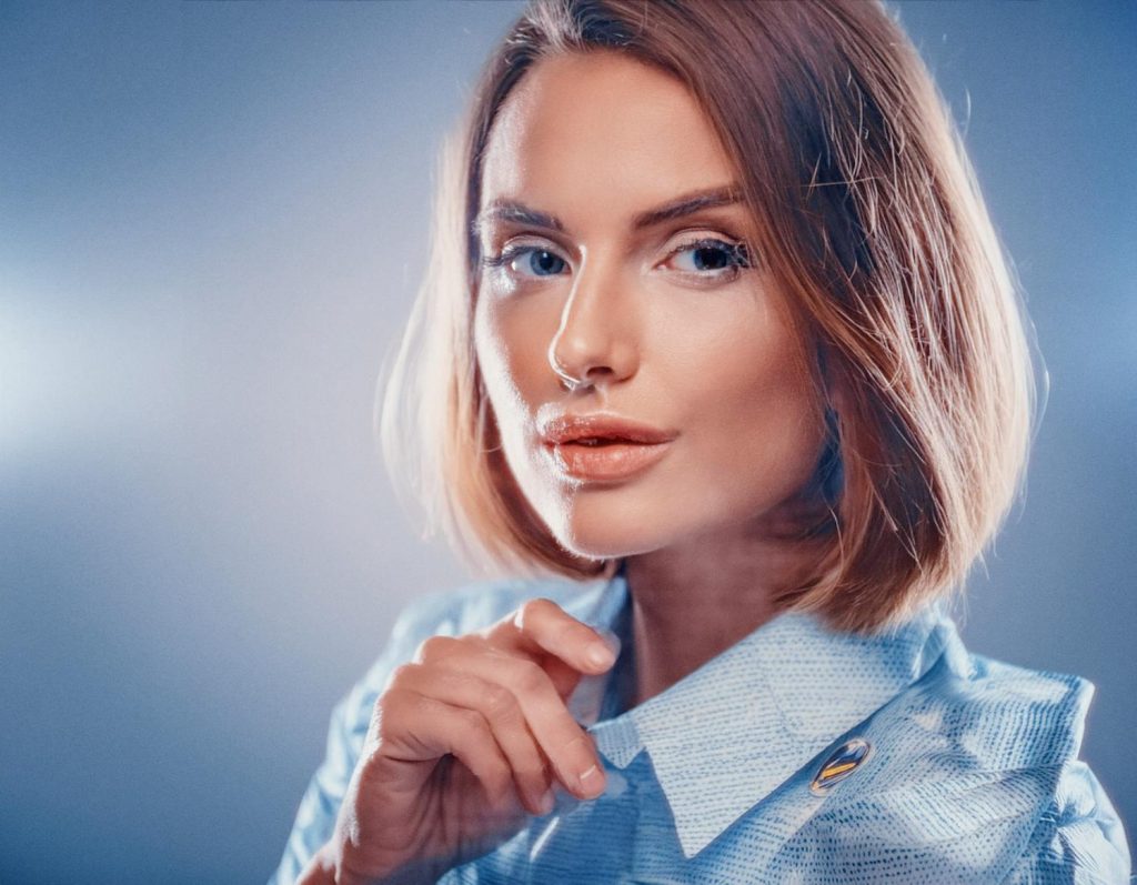 A woman with shoulder-length light brown hair and a neutral expression exudes timeless elegance in a light blue buttoned shirt, her hand gracefully holding the collar against a softly lit, blue-toned backdrop.