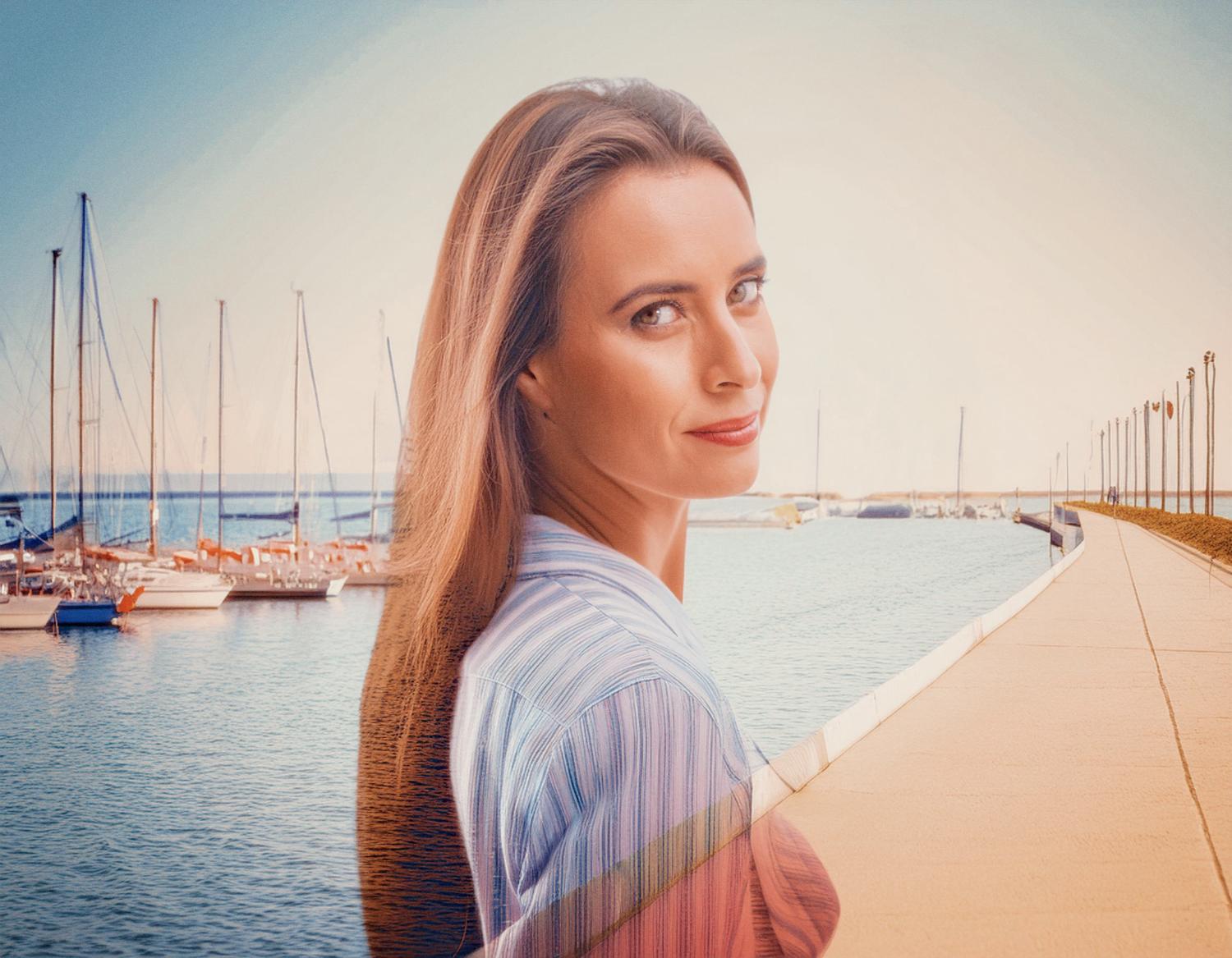 A stylish woman with flowing hair smiles confidently, her chic Osprey Packs bag over her shoulder. She stands by a serene marina under a clear blue sky.
