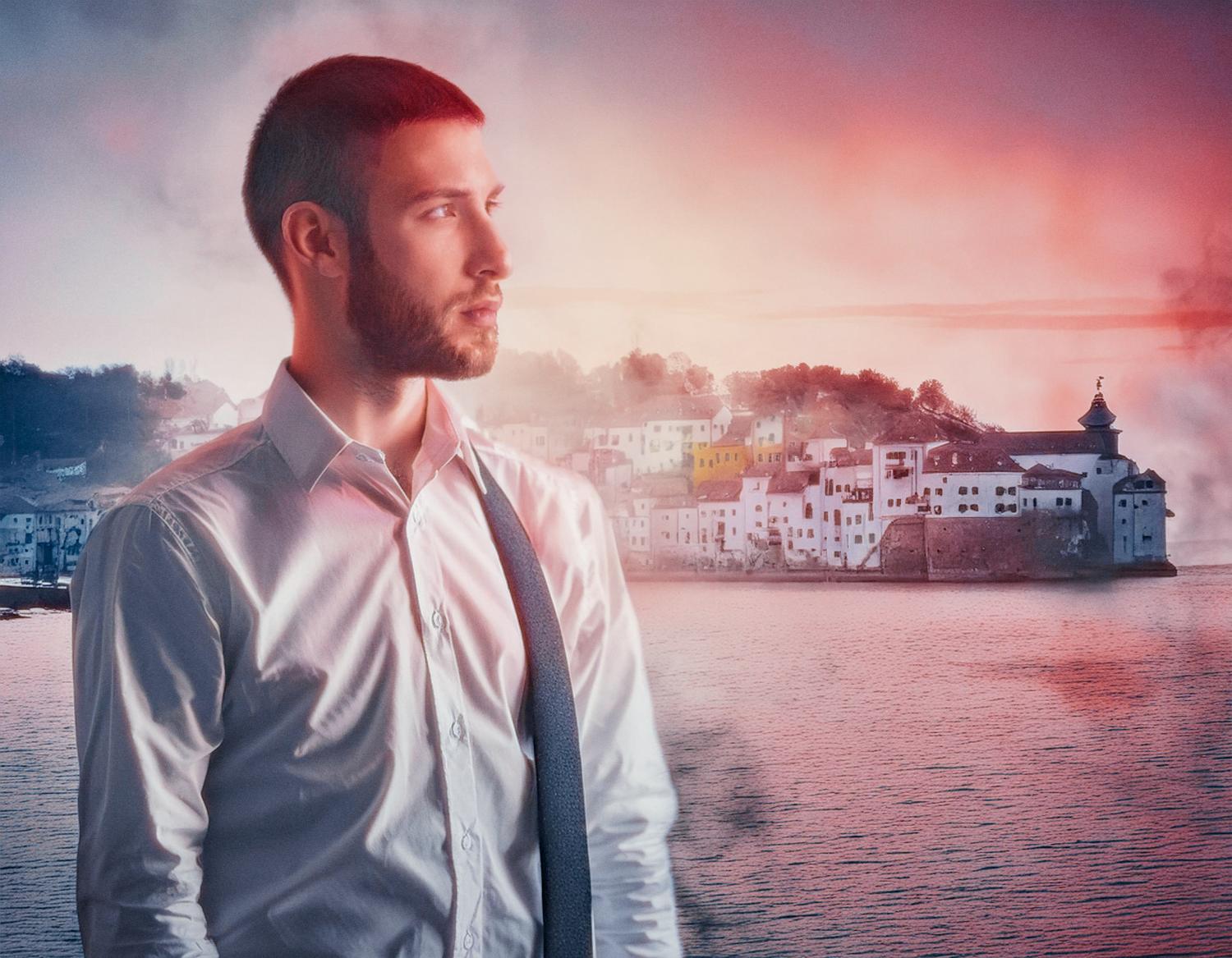 A bearded man with short hair in a crisp white shirt and dark tie, showcasing a stylish Timbuk2 Vapor Sling Crossbody Bag. He gazes to the side as a vibrant sunset casts a dreamy glow over the coastal town in the background. The soft lighting and mist add to the surreal, fashion-forward ambiance.