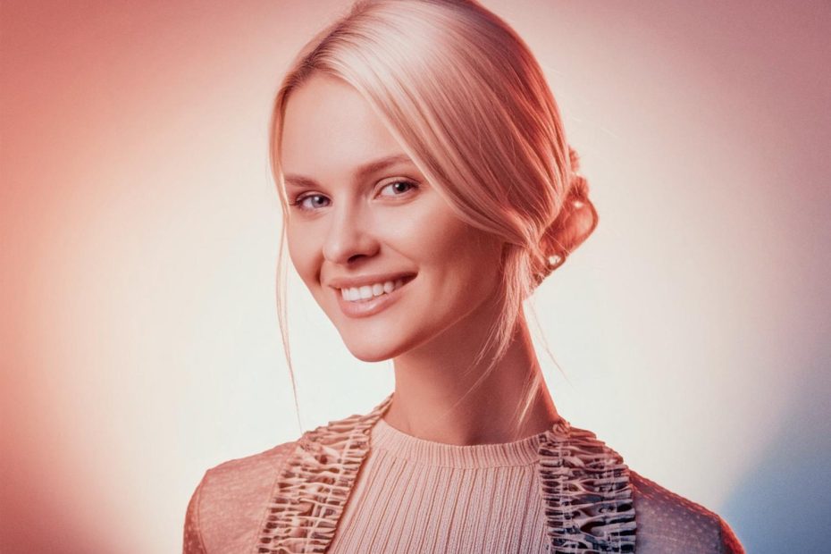 A woman with a sleek blonde low bun smiles warmly, her textured top and chic Timbuk2 Vapor Sling Crossbody Bag exuding effortless travel style. The gradient backdrop enhances the inviting aura.