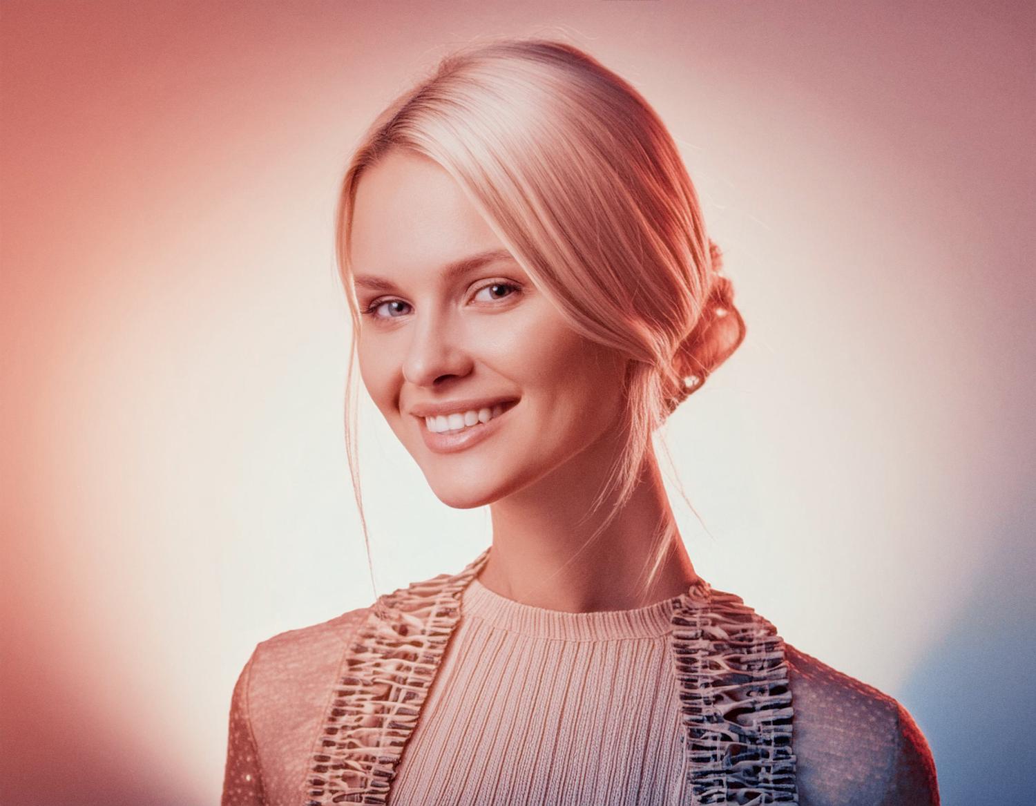 A woman with a sleek blonde low bun smiles warmly, her textured top and chic Timbuk2 Vapor Sling Crossbody Bag exuding effortless travel style. The gradient backdrop enhances the inviting aura.