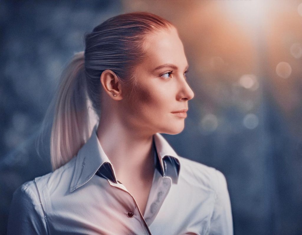 A stylish blonde woman with a sleek ponytail sports a chic light-colored collared shirt and a trendy Timbuk2 Vapor Sling bag, exuding calm travel vibes.