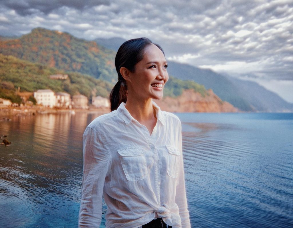 A woman in a chic white shirt smiles by calm waters, her stylish Heritage Waist Pack slung casually over her shoulder. Idyllic houses and lush green hills create the perfect serene backdrop.