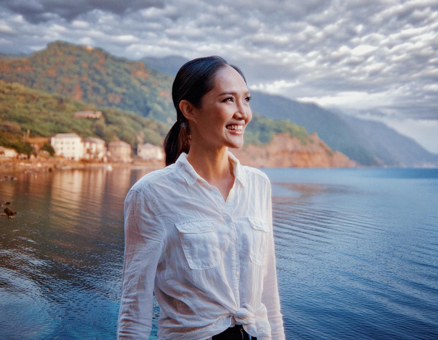 A woman in a chic white shirt smiles by calm waters, her stylish Heritage Waist Pack slung casually over her shoulder. Idyllic houses and lush green hills create the perfect serene backdrop.