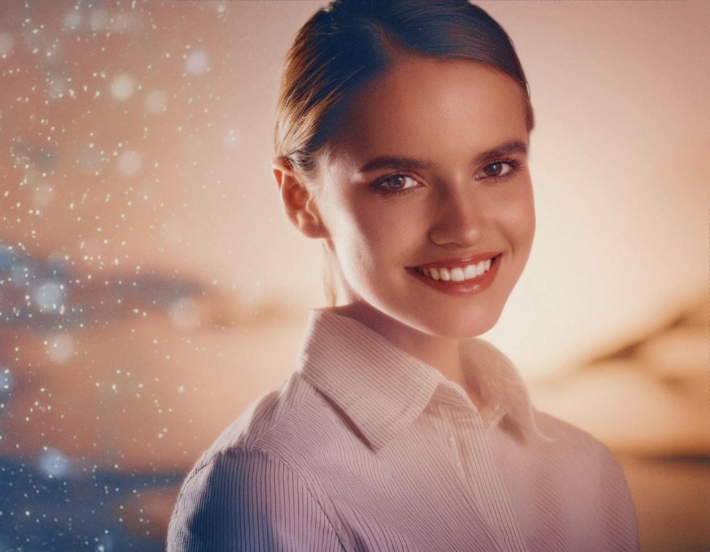 A stylish individual with dark hair pulled back smiles confidently at the camera in a chic, light-colored collared shirt. The dreamy, blurred background features warm and cool tones with sparkling particles, reminiscent of the Incase A.R.C. Collection aesthetic.