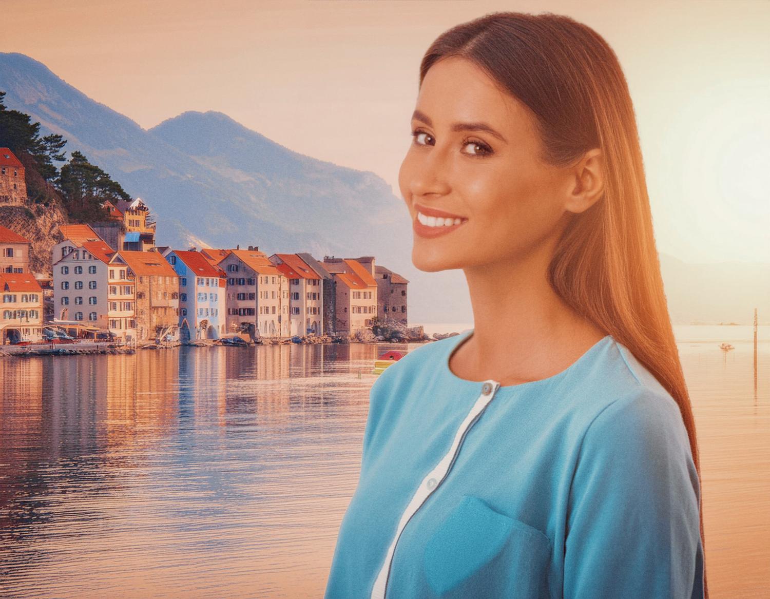 A woman with long brown hair, styled in a chic light blue top over a white shirt, smiles against the picturesque backdrop of a coastal town at sunset.