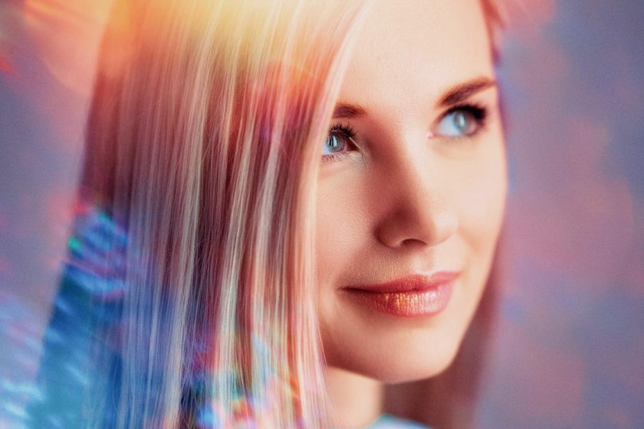 A blonde woman with soft blue eyes smiles gently. She sports a trendy Herschel Supply Roll Top Backpack and stylish headphones, surrounded by dreamy, colorful light effects.