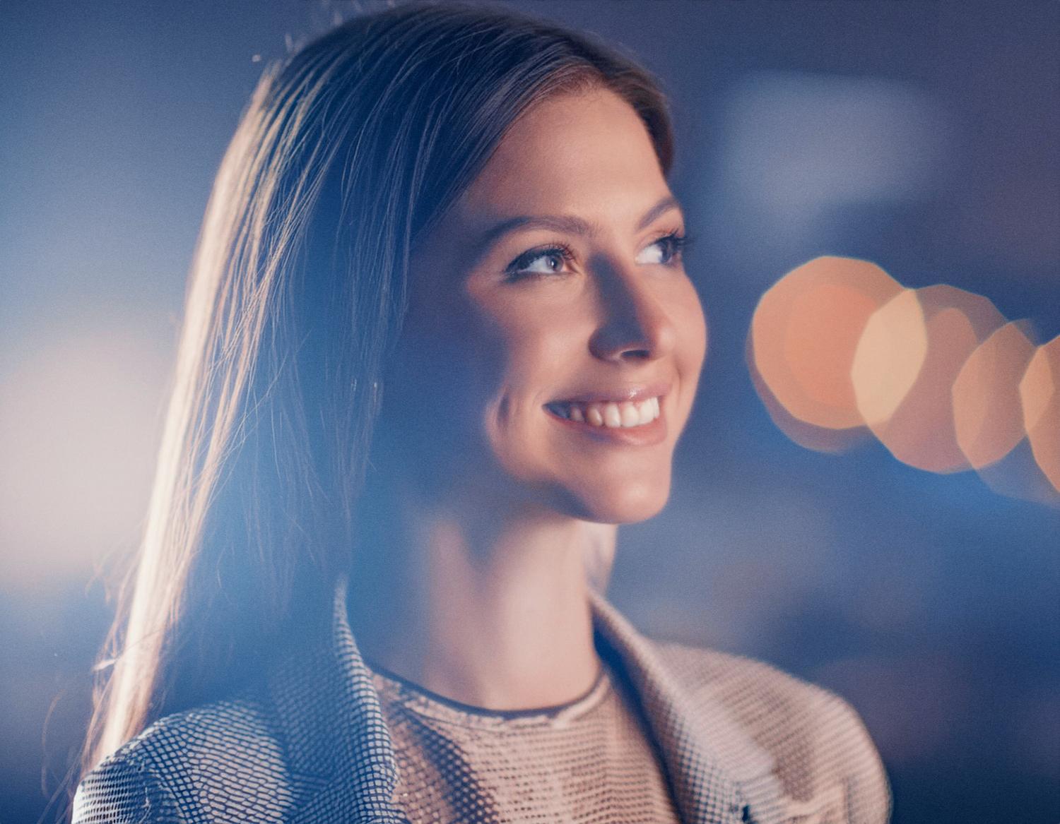 A woman with flowing brown hair beams in a checkered blazer and chic Herschel Roll Top Backpack. Soft bokeh lights set a stylish backdrop.