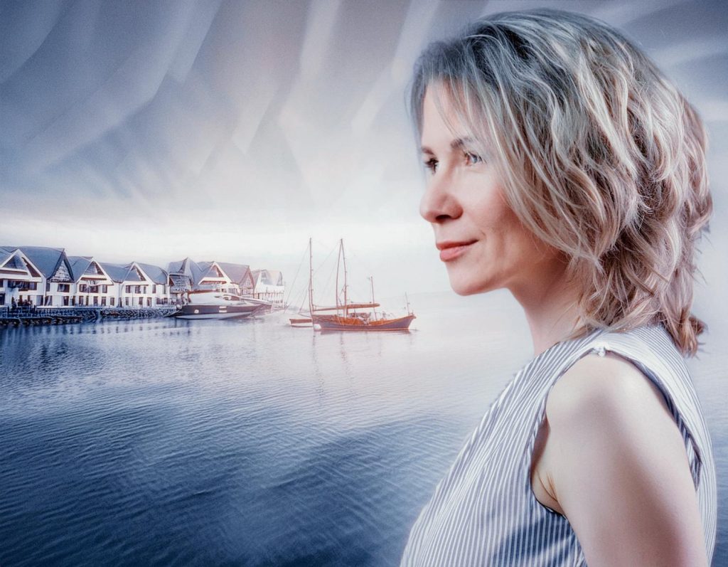 A reflective blonde woman in a chic striped top stands by tranquil waterfront with docked sailboats and stilt houses, complemented by her stylish Timbuk2 Spark Mini Backpack—a must-have accessory for the modern traveler.