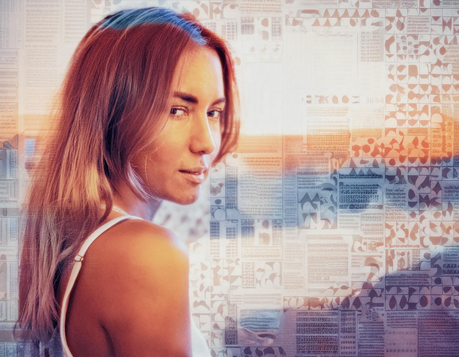 A woman with flowing auburn hair in a white tank top gazes over her shoulder, framed by geometric patterns and warm hues, exuding timeless elegance and sophisticated style.