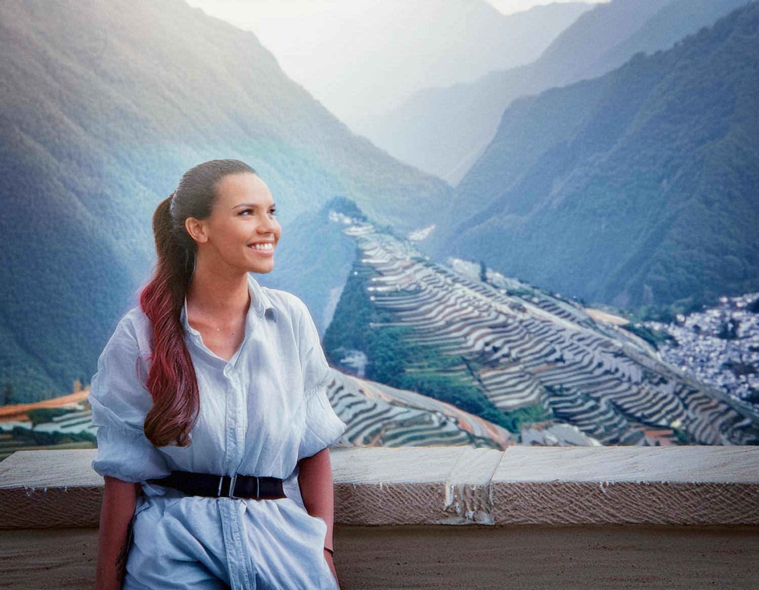 A smiling woman with a long ponytail poses amidst terraces in a chic light blue shirt and high-waisted pants, sporting a stylish Samsonite Brief backpack.
