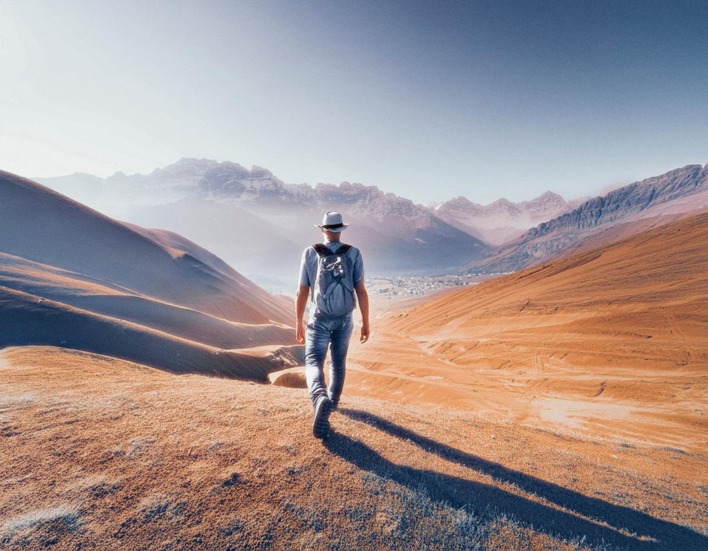A stylish traveler, with a chic hat and backpack, walks through an orange desert landscape. Misty mountains loom in the distance. Adventure calls. #HeritageVibes