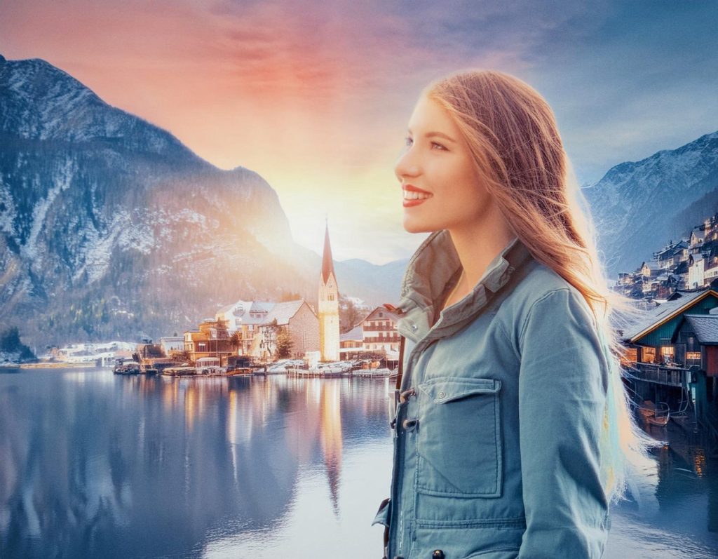 A stylish woman with long blonde hair in a chic blue jacket, smiles by a serene lake and picturesque village, her unique suitcase adding travel flair as the setting sun casts a warm glow.